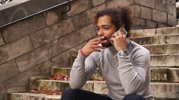 Schöner halb amerikanischer Kerl telefoniert mit dem Smartphone und raucht auf Stufen. — Stockvideo