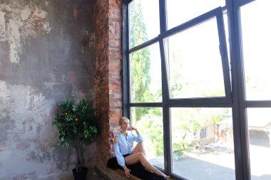 Çekici genç kadın gülümseyerek poz veriyor ve pencere pervazına üzerinde oturur