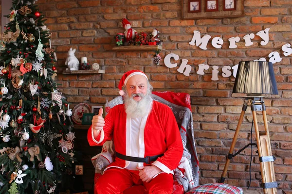 Der energiegeladene Weihnachtsmann hebt den Daumen, sitzt auf einem Sessel im Wohnzimmer. — Stockfoto