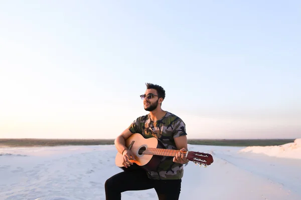 Handsome Arabian guy playing guitar, standing on hill among sand