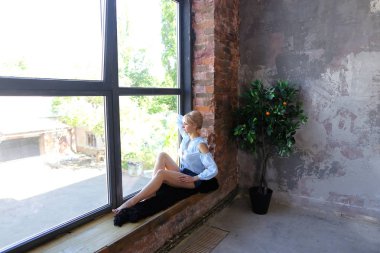 Çekici genç kadın gülümseyerek poz veriyor ve pencere pervazına üzerinde oturur