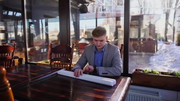 Arquitecto esperando al cliente en la cafetería con el proyecto de arquitectura roll y mirando el reloj inteligente — Vídeos de Stock