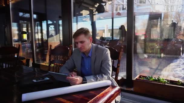 Инженер просматривает планшет и сидит в кафе со смартфоном и ролл-проектом на столе . — стоковое видео