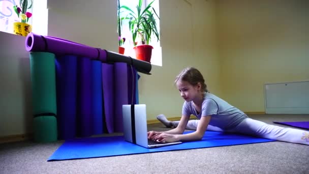 Liten flicka på amerikanska utseende sitter på golvet i cross-twist position och spelar på laptop. — Stockvideo