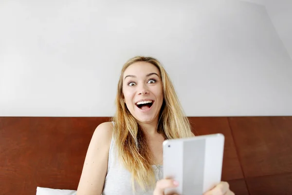 Młoda kobieta szczęśliwa wstrząśnięty z tabletu, patrząc na kamery w białe łóżko — Zdjęcie stockowe