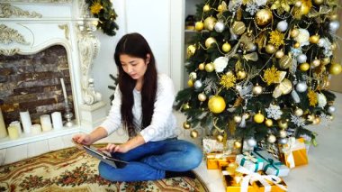 Sevimli kadın Online rezervasyon veya rezervasyon tatil için tablet kullanarak öznitelikleri ve arka plan Noel ağacı ve parlak odasında şömine oturur.