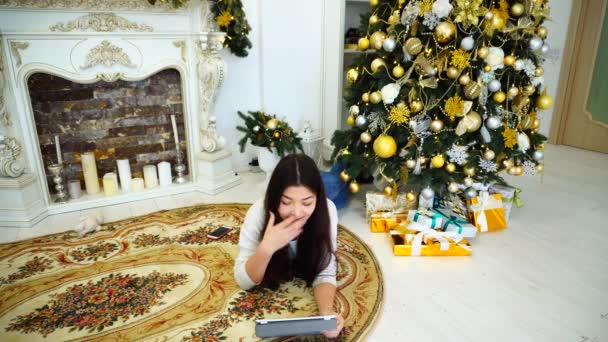 Mujer de negocios en vísperas de las vacaciones a través de la tableta termina todos los asuntos de negocios en casa acostado sobre una alfombra en el fondo Árbol de Navidad y chimenea en la habitación . — Vídeo de stock