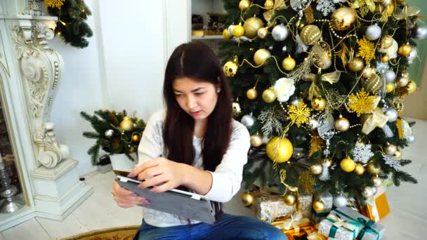 Roztomilá žena pomocí Tablet pro on-line rezervace nebo rezervace dovolené atributy a sedí na pozadí vánoční strom a ve světlé místnosti krb. — Stock video