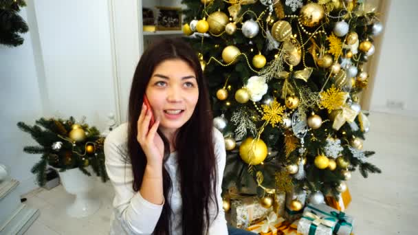 Splendida ragazza che parla al telefono e ordina regali, si siede su sfondo decorato albero di Natale e camino in camera luminosa . — Video Stock