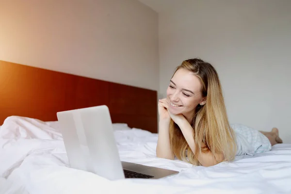 Mujer joven mirando el ordenador portátil que trabaja en casa de forma remota en la cama blanca — Foto de Stock