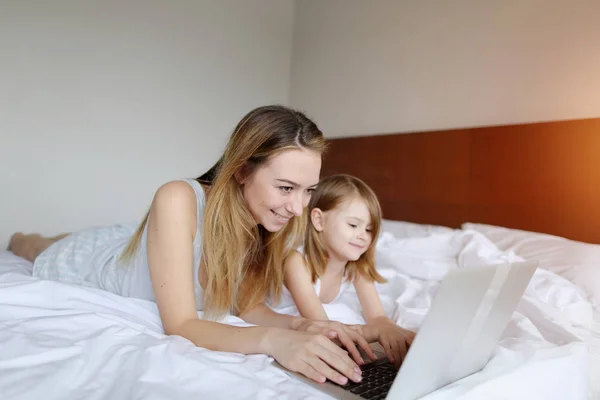 Mère femme et petite fille utilisent ordinateur portable ensemble au lit blanc avec soleil — Photo