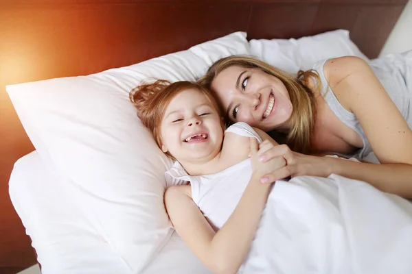 Beleza mãe abraços pequena filha no branco cama com sol — Fotografia de Stock