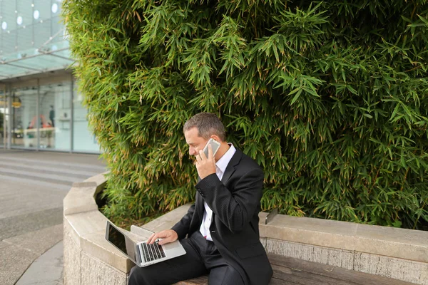 Manager talking by smartphone and working with laptop outside in