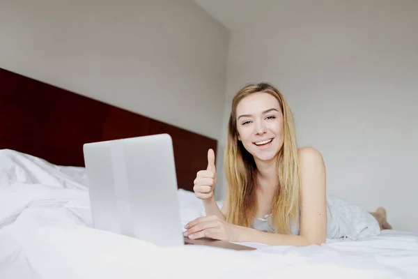 Joven mujer feliz espectáculo como pulgares hacia arriba con el ordenador portátil de trabajo a distancia en la cama blanca — Foto de Stock