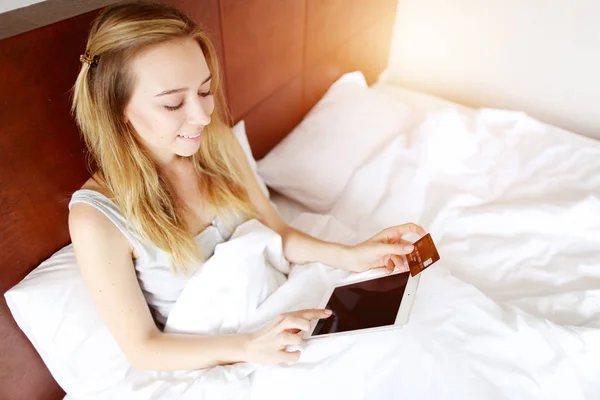 여행에 대 한 돈을 지불 하는 젊은 여자 보유 신용 카드 직불 카드와 집에서 햇빛으로 흰 침대에 누워 태블릿 — 스톡 사진