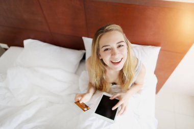genç kadın banka kredi kartı tutan ve yatakta beyaz evde güneş ışığı ile döşeme kamera tablet bakar