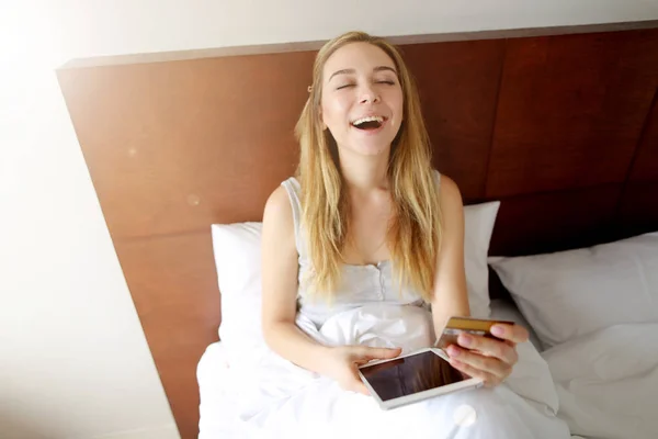super happy woman with credit card and tablet laughing in white bed