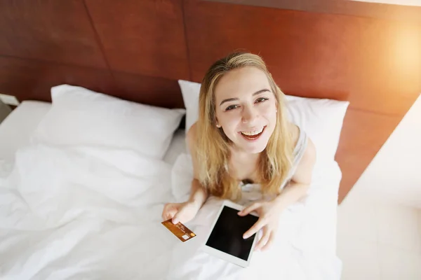 young woman holds debit credit card and tablet looks at camera laying in white bed at home with sunshine