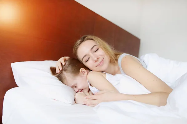 Ibu cantik mengantuk memeluk putri kecil di tempat tidur putih dengan sinar matahari — Stok Foto