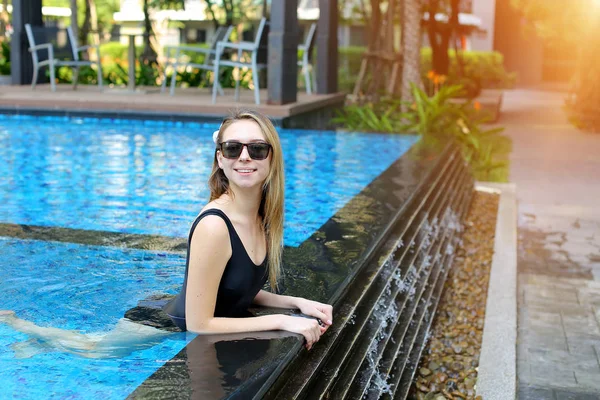 Joven hembra mirando a la cámara con gafas de sol disfrutar de la natación en la piscina azul al aire libre, relajarse cerca de las palmas y edificio de apartamentos en el país tropical — Foto de Stock
