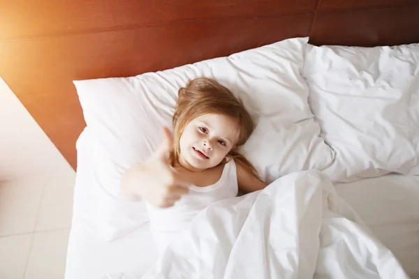 Портрет маленькой дочери показывает большие пальцы вверх, как улыбка лежа на белой кровати дома на солнечное доброе утро — стоковое фото