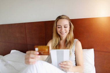 güneş ışığı ile beyaz yatakta kredi kartı ve tablet tarafından yeni ev satın kadın odaklanmak