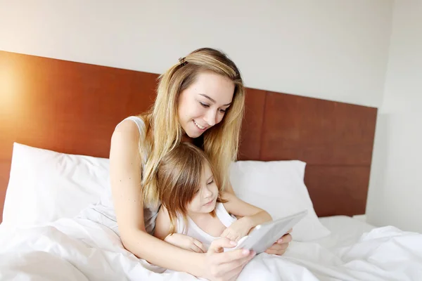 Mãe e filha usam tablet na cama branca com sol — Fotografia de Stock