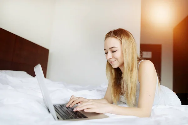 Mujer joven feliz con el ordenador portátil trabajando en casa de forma remota en la cama blanca — Foto de Stock