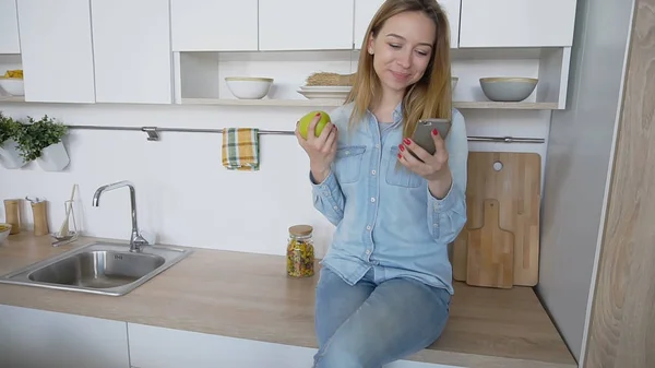 Muchacha atractiva utiliza el teléfono inteligente y come manzana, sentado en kitc — Foto de Stock