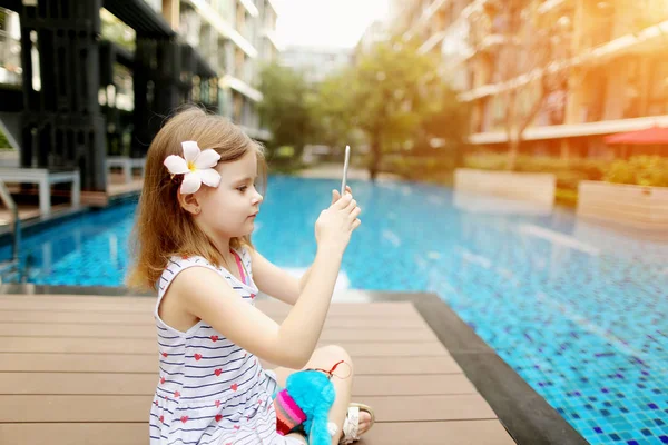 Niña hacer foto en la tableta sentado cerca de la piscina con flores en el pelo en el día soleado — Foto de Stock