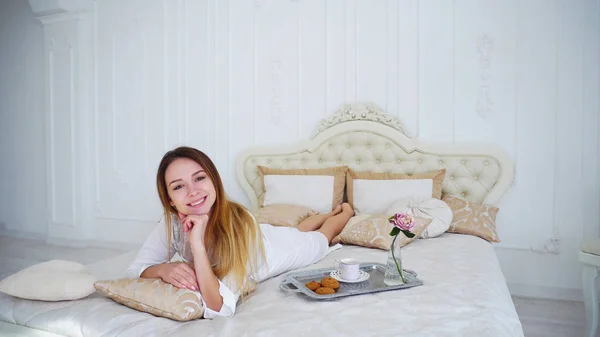 Chica modelo sonriendo en la cámara acostado en la cama en el dormitorio brillante . — Foto de Stock