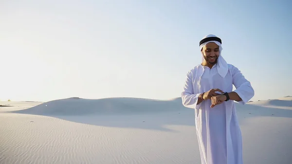 Modern male sheikh uses smart watch and stands in middle of bott