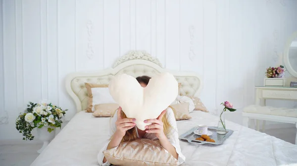 Retrato de linda hembra sosteniendo almohada-corazón, posando y sonriendo mientras miente — Foto de Stock