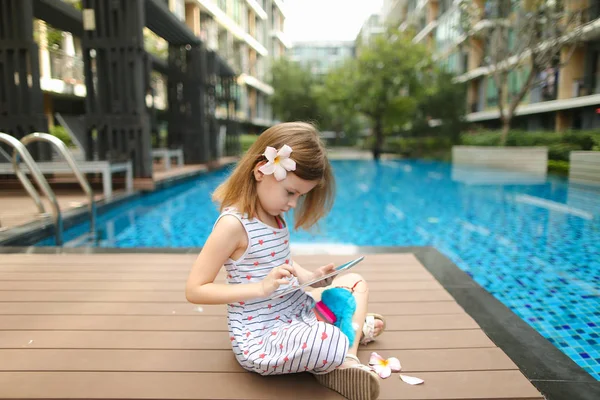 Niña utiliza la tableta sentada cerca de la piscina usando f — Foto de Stock