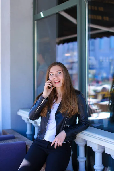 Cute female person speaking by smartphone near window of street cafe.