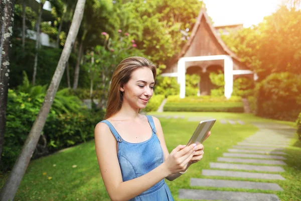 Retrato del sol de la mujer joven usando tableta y sonriendo en las palmas verdes y fondo de la casa en Tailandia — Foto de Stock