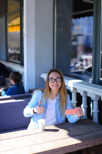 Chica mostrando los pulgares hacia arriba, sentado en la cafetería de la calle con teléfono inteligente . — Foto de Stock