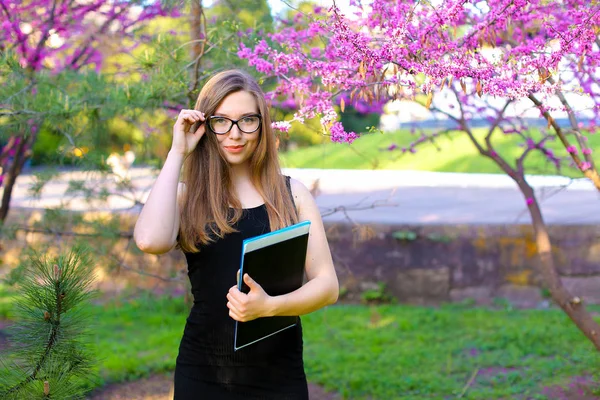 Studentessa toccando bicchieri in giardino e in piedi con documenti in cartella nera . — Foto Stock