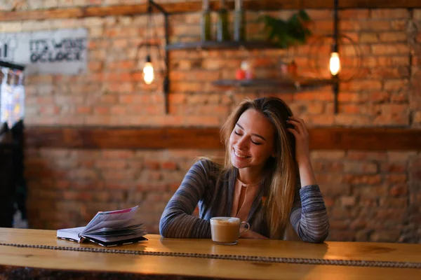 Piękna dziewczyna siedzi w pobliżu tabeli filiżankę cappuccino w przytulnej kawiarni. — Zdjęcie stockowe