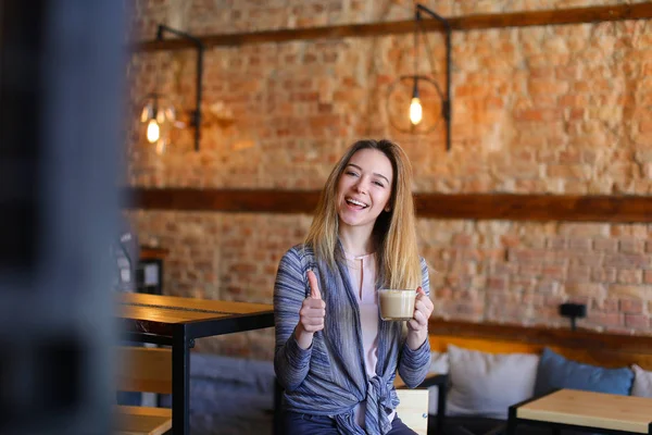 Zufriedene Mädchen am Tisch mit einer Tasse Cappuccino in einem gemütlichen Café mit ungewöhnlichem Interieur. — Stockfoto