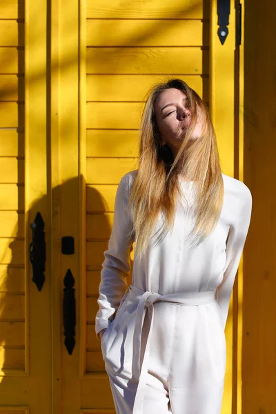 Menina europeia bonita vestindo terno branco em pé perto da porta amarela em raios de sol . — Fotografia de Stock