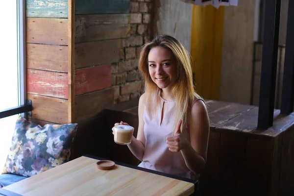 Hübsches Mädchen sitzt auf Stuhl neben Holztisch mit einer Tasse Cappuccino in der Mittagspause. — Stockfoto