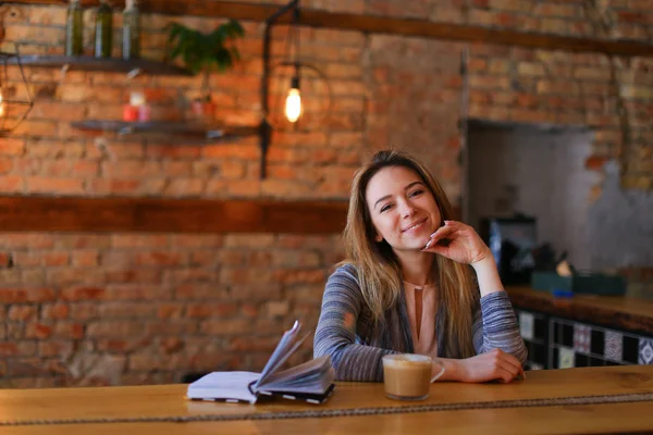 Szczęśliwa kobieta siedzi w pobliżu tabeli filiżankę cappuccino i notebook w przytulnej kawiarni z niezwykłym wnętrzu. — Zdjęcie stockowe