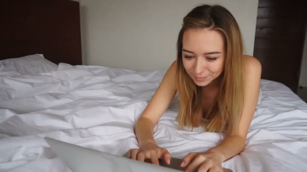 Bella ragazza sdraiata a letto e chatta nel computer portatile . — Video Stock