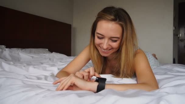 Persona bastante femenina acostada en la cama con edredón blanco y usando reloj inteligente . — Vídeos de Stock