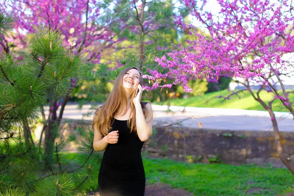 Mujer joven hablando por teléfono inteligente en el parque cerca de los árboles en flor . —  Fotos de Stock