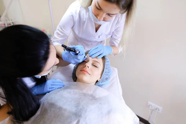 Cosmetologist doing permanent eyebrow makeup for woman at cabinet with nurse.