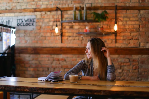 Nettes Mädchen sitzt am Tisch mit einer Tasse Cappuccino und Notizbuch in gemütlichem Luncheonette. — Stockfoto