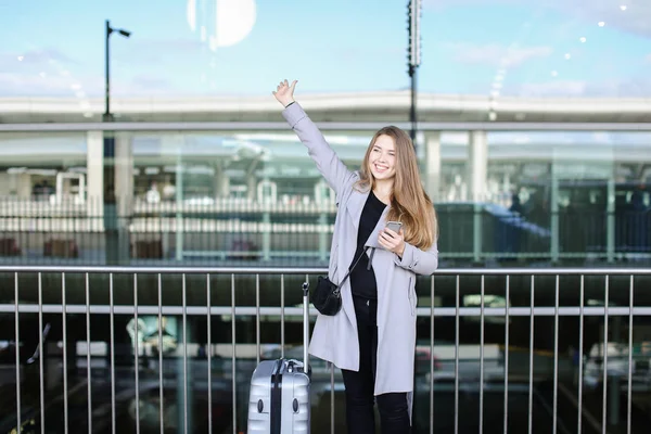 Caucasian female person stopping taxi with raised hand, valise and smartpone near airport. — Stock Photo, Image