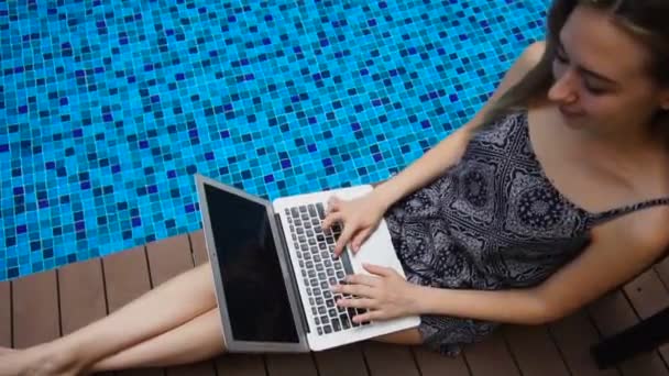 Jeune femme couchée près de la piscine bleue et utilisant un ordinateur portable au ralenti . — Video